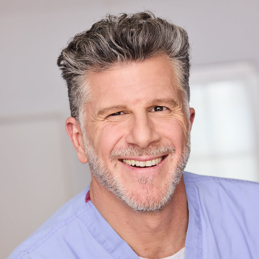 Professional headshot of a smiling healthcare provider wearing light blue medical scrubs in a clinical setting